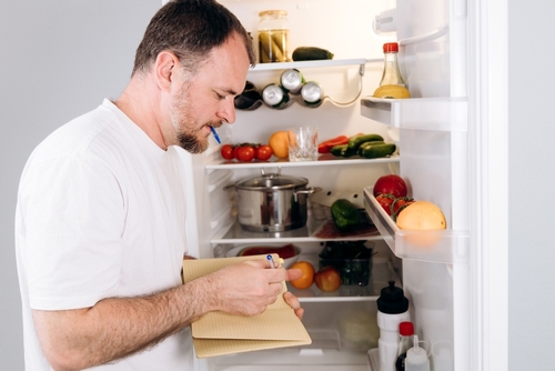 homem branco anotando itens em um papel de frente para uma geladeira aberta - Assaí Atacadista - dicas de cozinha para quem mora sozinho
