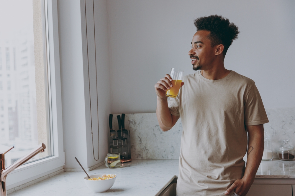 jovem negro feliz tomando suco na cozinha - hábitos para praticar - Assaí Atacadista