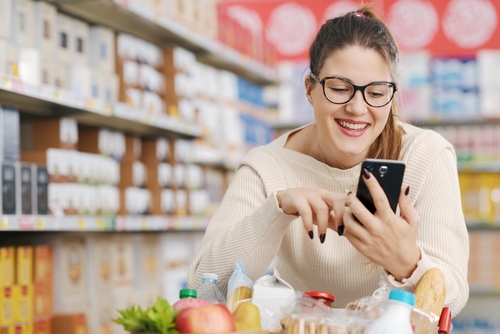 mulher branca feliz usando o celular no mercado - App Meu Assaí 2024 - Assaí Atacadista