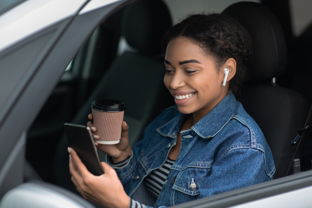 mulher negra sorrindo segurando um celular sentada em um carro - tecnologia para economizar - Assaí Atacadista