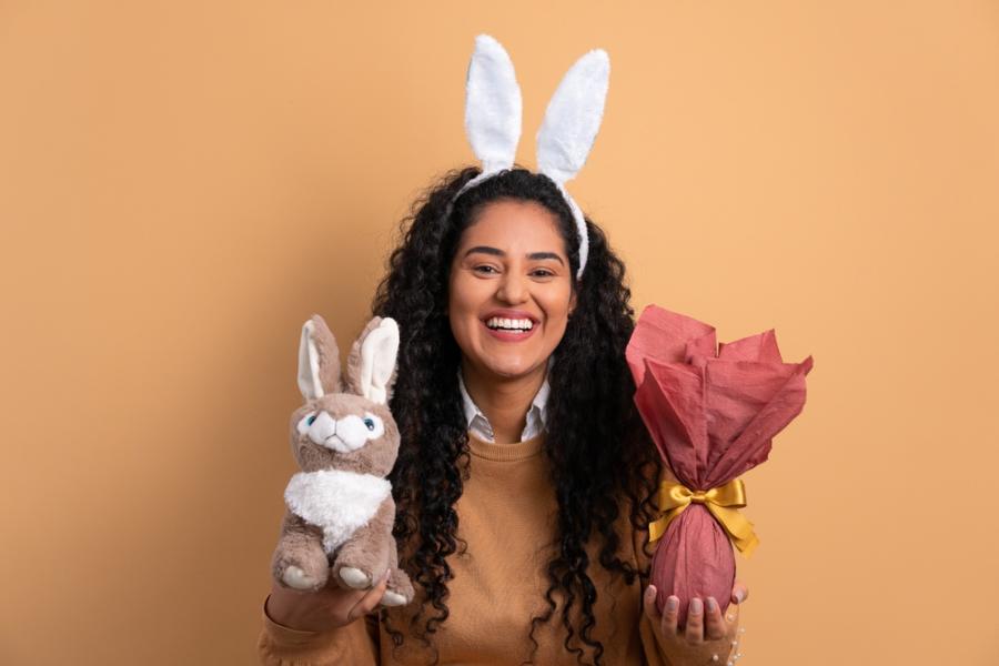 mulher sorridente com orelhas de coelho segurando um coelho de pelucia e um ovo de chocolate nas mãos - presente para o dia da mulher - assaí atacadista